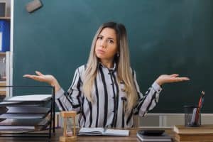 Trump administration cuts Harvard federal funding: young woman teacher sitting at school desk in front of blackboard in classroom checking homework of students looking disappointed spreading arms to the sides